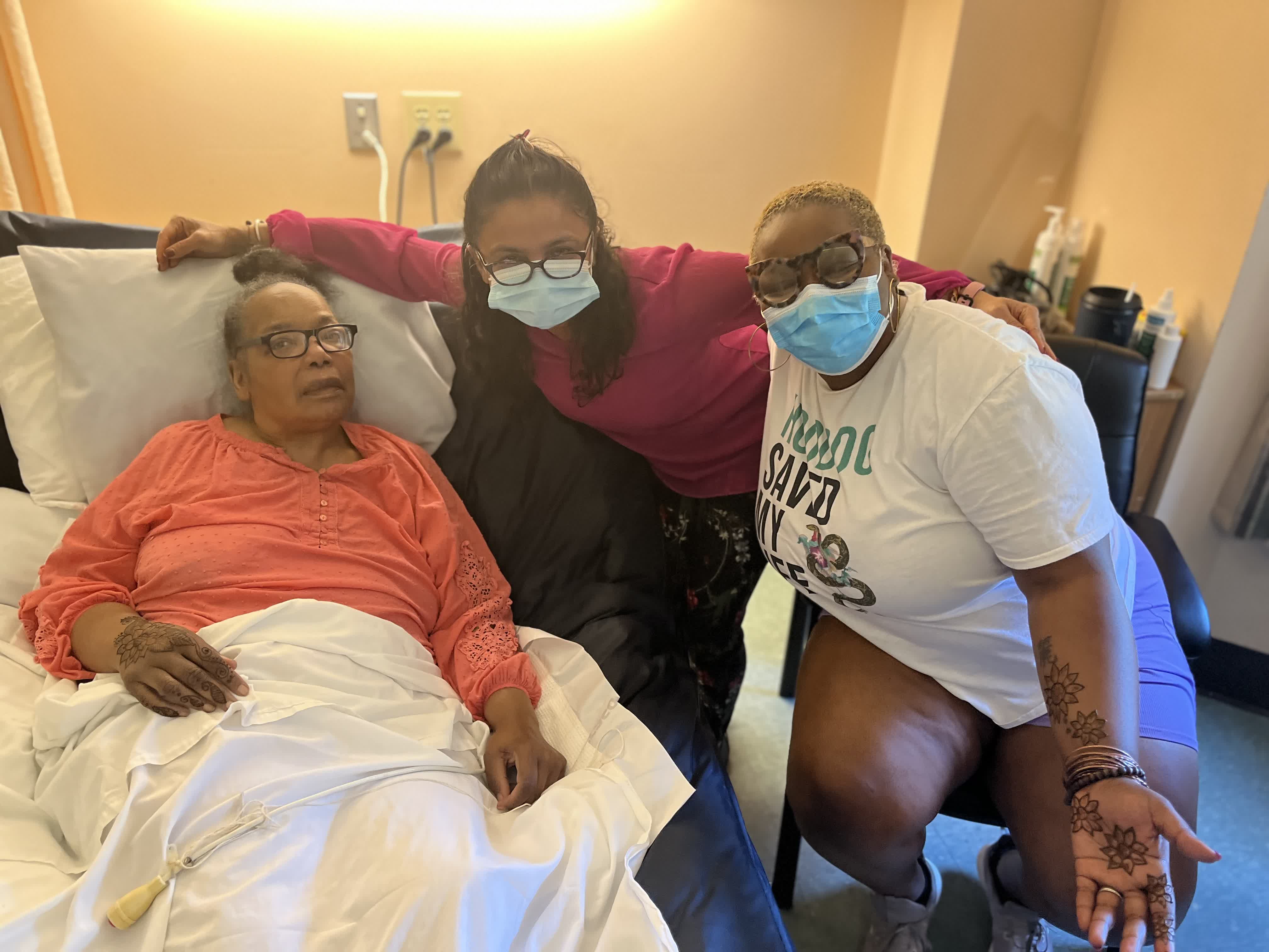 A hospice patient with fresh henna on her hand, smiling with two family visitors by her bedside